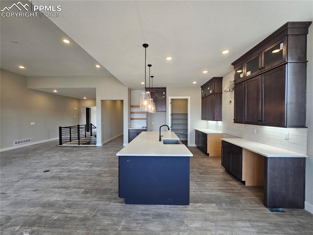 Kitchen with an island with sink, decorative backsplash, glass insert cabinets, dark brown cabinetry, and pendant lighting