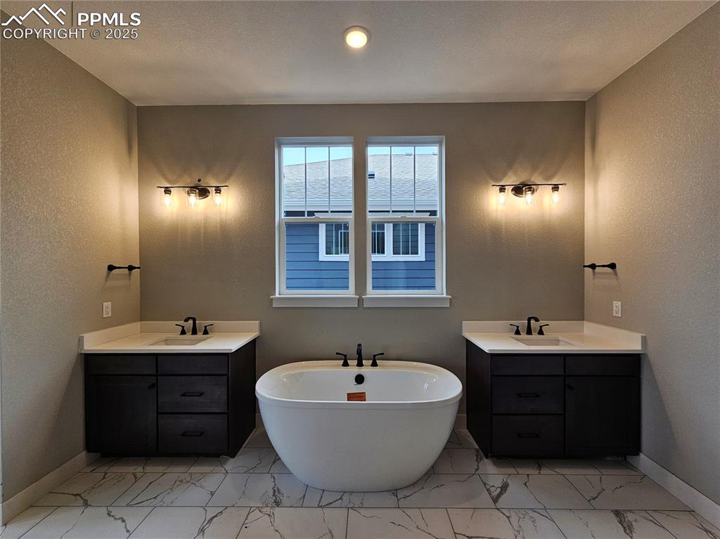 Bathroom with a textured wall, two vanities, a soaking tub, and light marble finish flooring