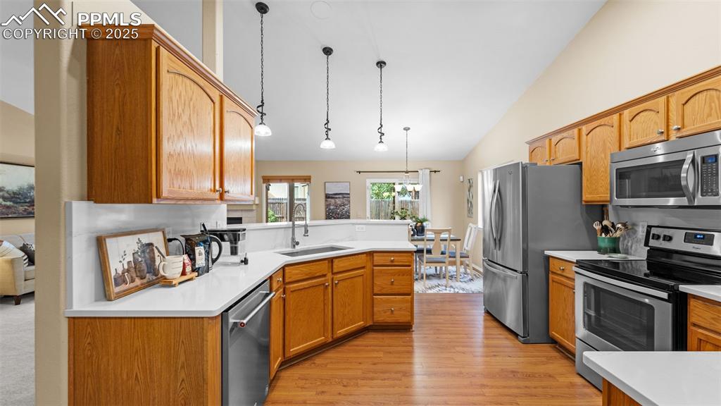 Kitchen featuring stainless steel appliances, decorative light fixtures, light wood finished floors, brown cabinetry, and high vaulted ceiling