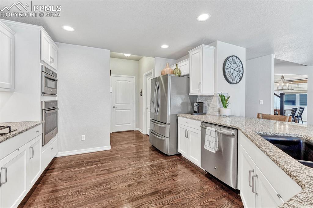 Kitchen with backsplash, white cabinetry, stainless steel appliances, light stone countertops, and dark wood-style flooring