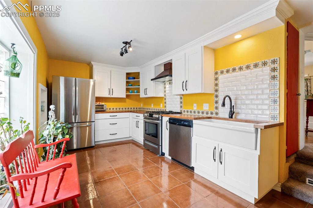 Kitchen featuring white cabinetry, appliances with stainless steel finishes, decorative backsplash, light tile patterned flooring, and wall chimney exhaust hood