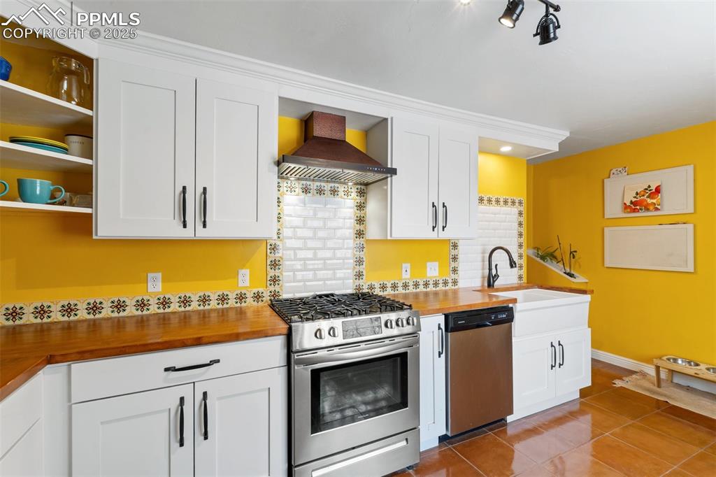 Kitchen featuring appliances with stainless steel finishes, white cabinetry, decorative backsplash, and wall chimney range hood