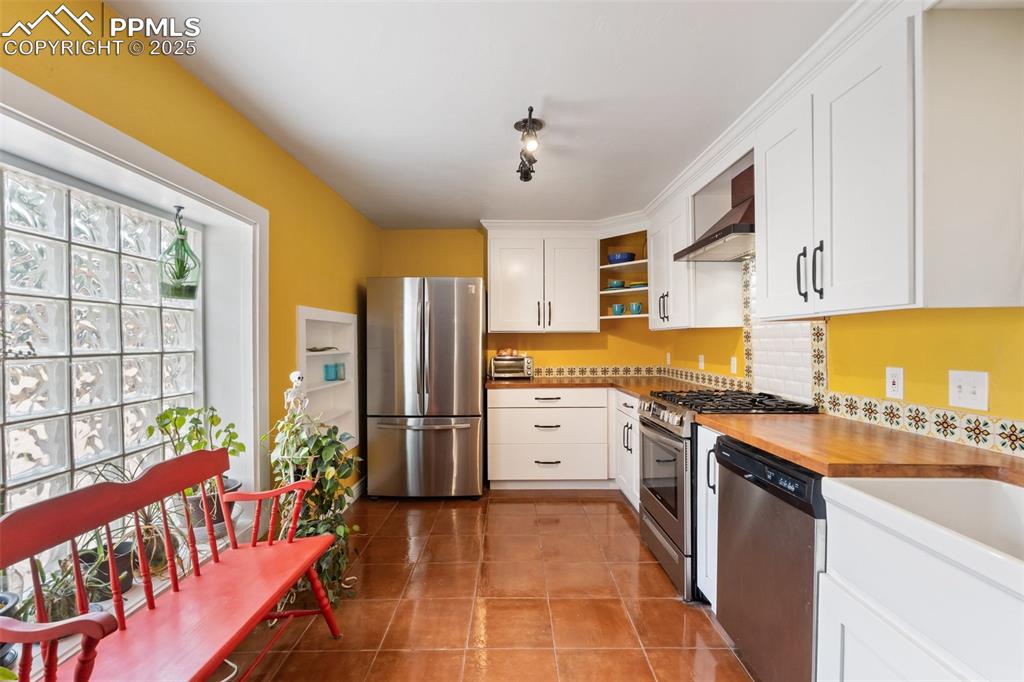 Kitchen with appliances with stainless steel finishes, white cabinetry, open shelves, wall chimney range hood, and light tile patterned floors