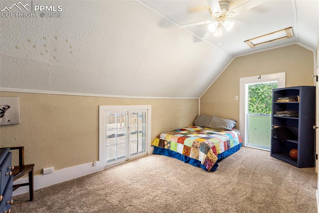 Carpeted bedroom with attic access, a textured wall, vaulted ceiling, access to exterior, and a ceiling fan