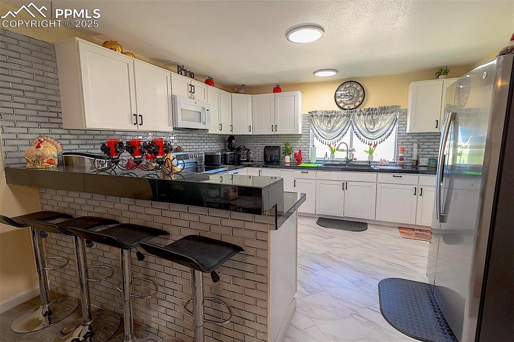 Kitchen with white microwave, freestanding refrigerator, light marble finish flooring, a peninsula, and a sink