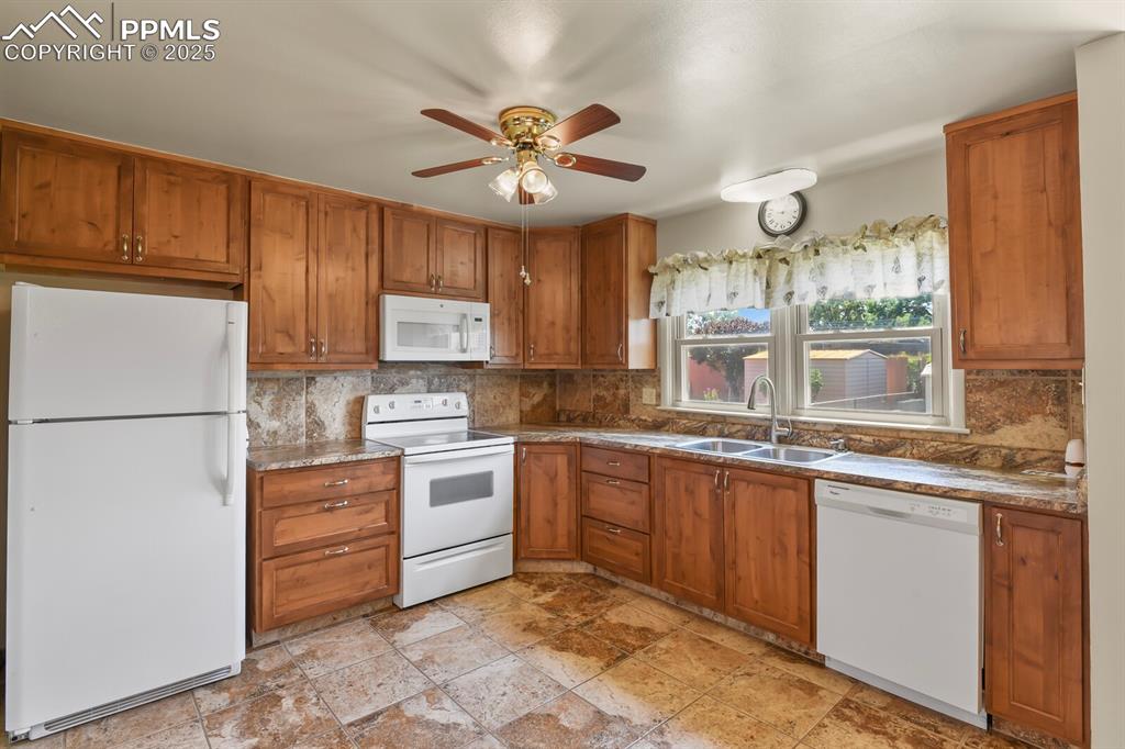 Kitchen featuring white appliances, solid wood cabinetry, backsplash, ceiling fan, and light stone tile flooring