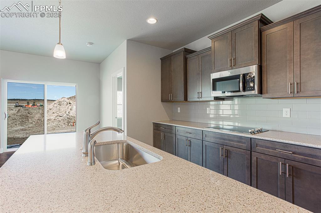 Kitchen with dark brown cabinets, light stone counters, stainless steel microwave, decorative backsplash, and decorative light fixtures