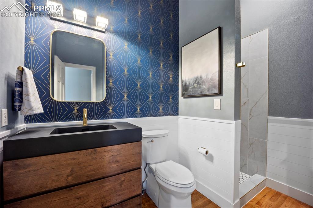 Full bathroom with wallpapered walls, toilet, wood finished floors, vanity, and wainscoting