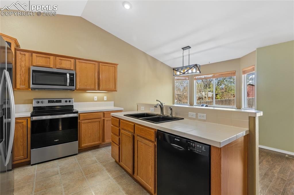 Kitchen featuring stainless steel appliances, wood finish cabinets, vaulted ceiling, hanging light fixtures, and tile counters