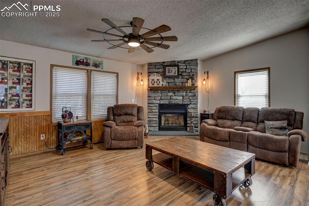 Living room featuring a textured ceiling, a stone fireplace, wooden walls, light wood-style flooring, and a ceiling fan