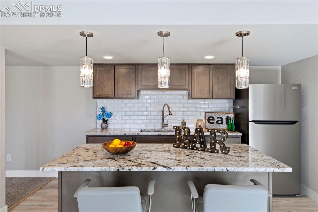 Basement Kitchenette/Bar with freestanding refrigerator, a center island with sink, a breakfast bar, light stone countertops, and light wood-style floors
