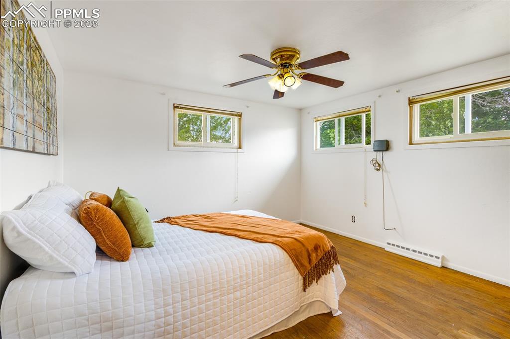 Bedroom with wood finished floors, multiple windows, a baseboard heating unit, and ceiling fan
