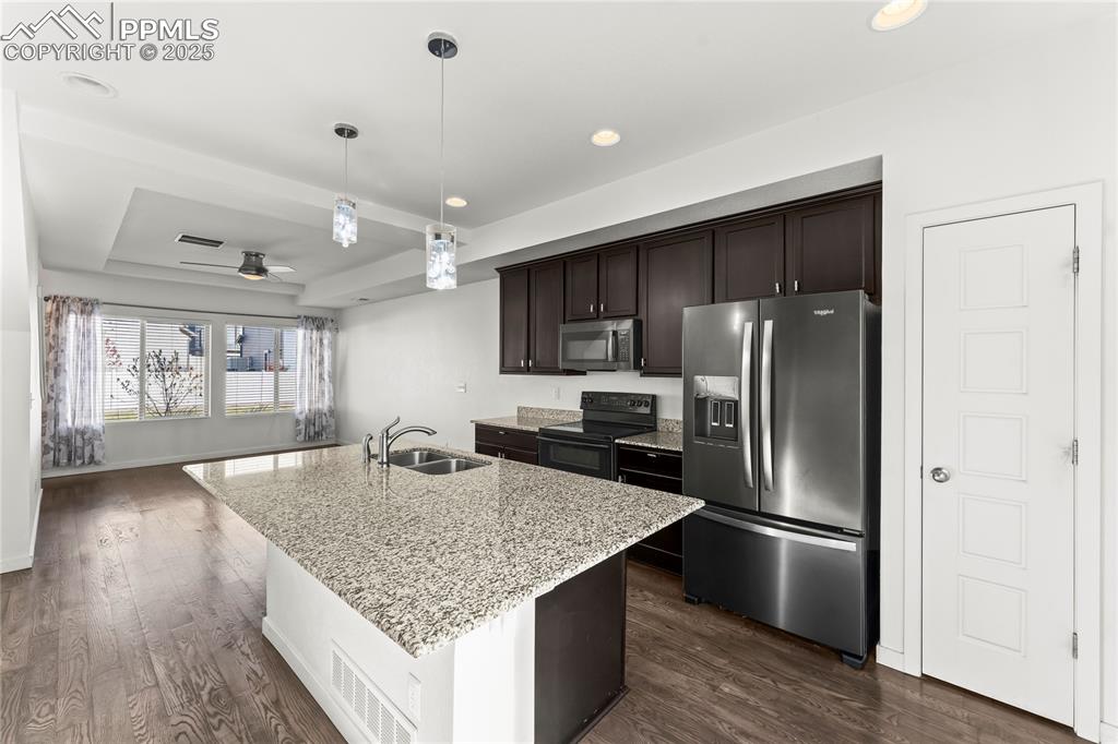 Kitchen with stainless steel appliances, dark brown cabinetry, light stone countertops, dark wood finished floors, and an island with sink