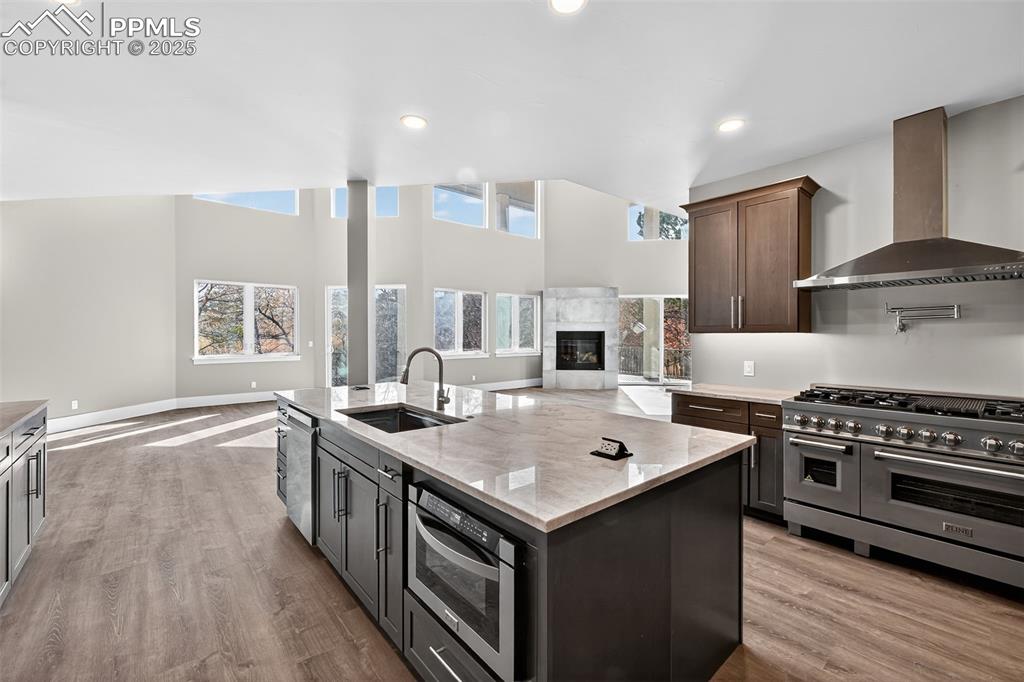 Kitchen featuring open floor plan, wall chimney range hood, stainless steel appliances, a kitchen island with sink, and a glass covered fireplace