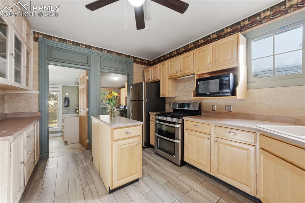 Kitchen with appliances with stainless steel finishes, light countertops, glass insert cabinets, light brown cabinets, and a ceiling fan