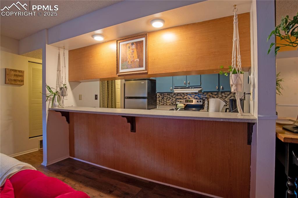 Kitchen featuring blue cabinetry, stainless steel appliances, tasteful backsplash, dark wood finished floors, and a kitchen bar