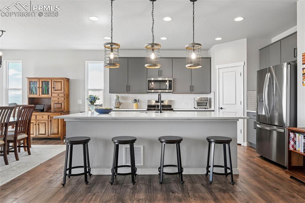 Kitchen with a center island with sink, stainless steel appliances, gray cabinets, a breakfast bar, and dark wood-style floors