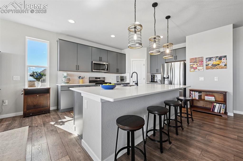 Kitchen featuring gray cabinets, a breakfast bar, an island with sink, decorative light fixtures, and dark wood-style floors