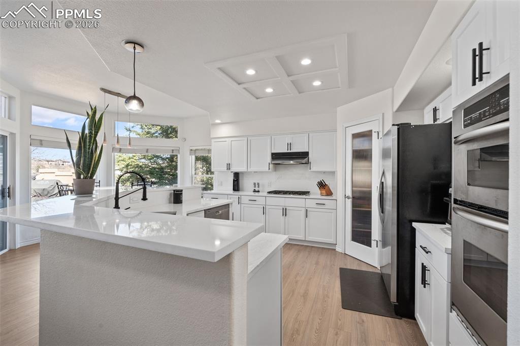 Kitchen featuring white cabinets, a large island with sink, stainless steel appliances, light wood-style floors, and hanging light fixtures