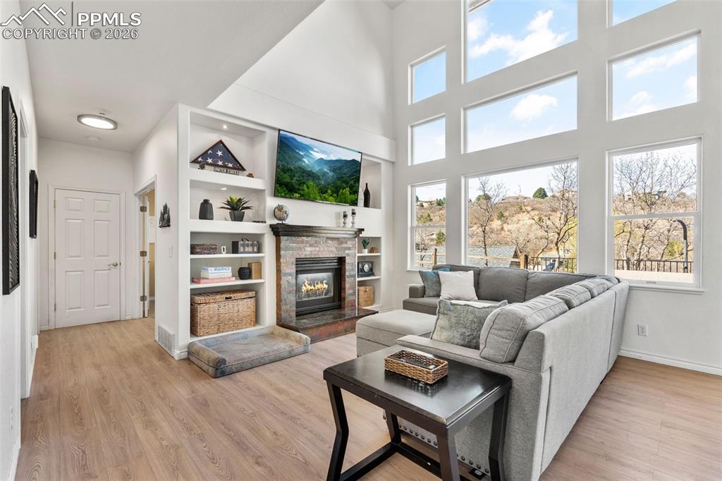 Living room with built in features, a stone fireplace, light wood-type flooring, and a high ceiling