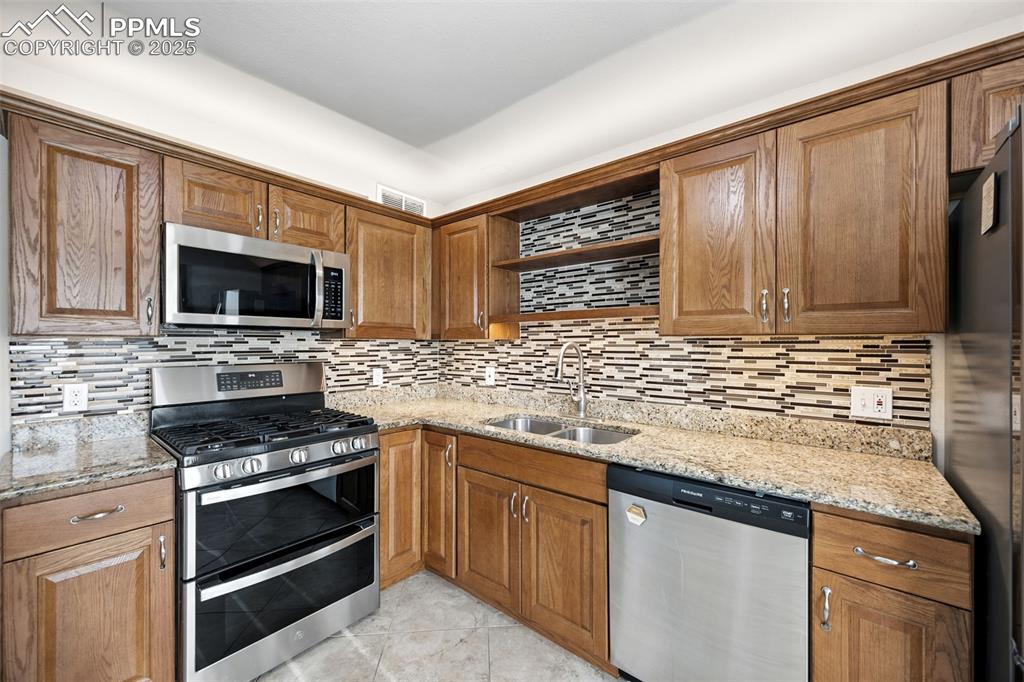 Kitchen with appliances with stainless steel finishes, light stone countertops, brown cabinetry, decorative backsplash, and light tile patterned flooring