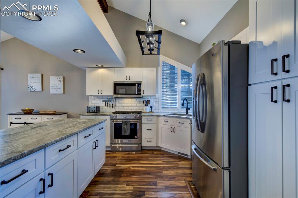 Kitchen with lofted ceiling, stainless steel appliances, white cabinetry, dark wood-style flooring, and light stone countertops