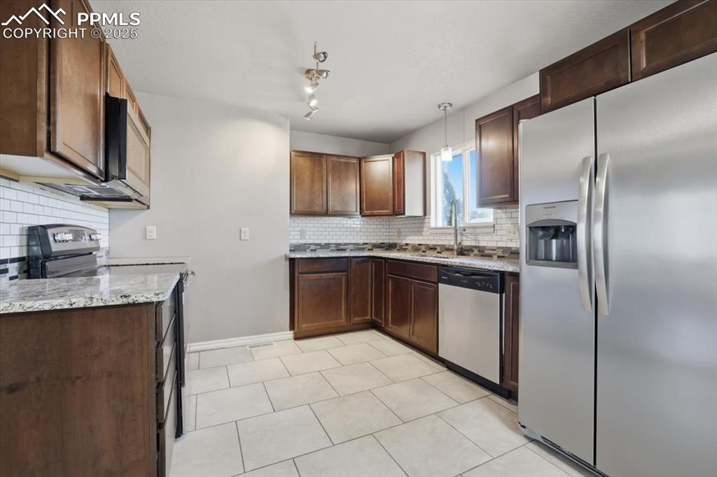 Kitchen featuring stainless steel appliances, light stone countertops, decorative backsplash, dark brown cabinets, and light tile patterned floors