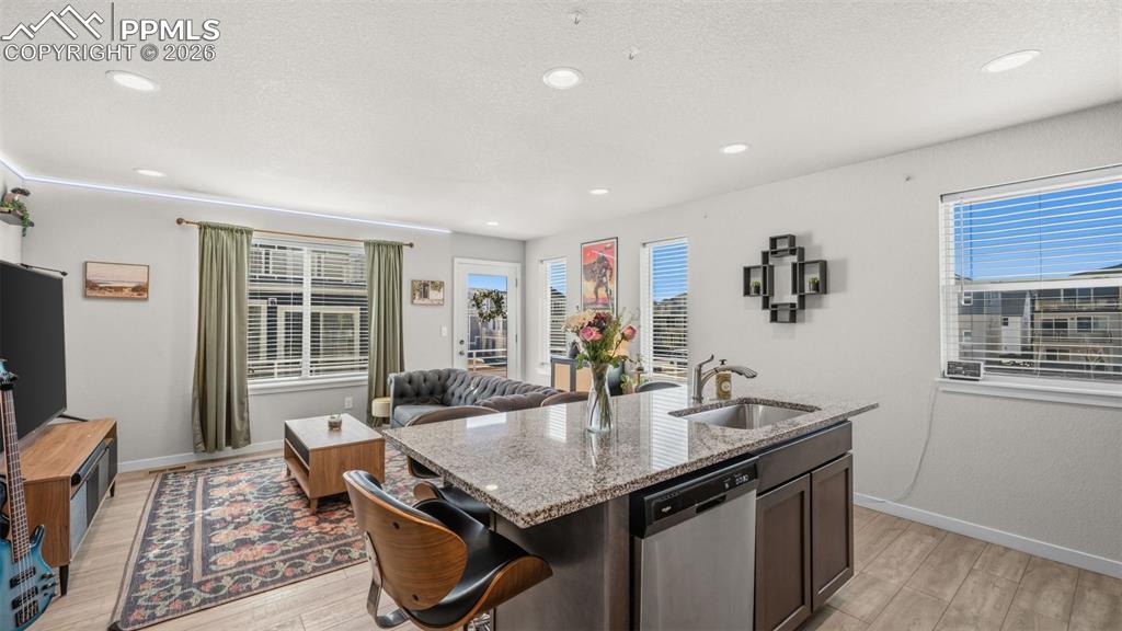 Kitchen featuring light stone countertops, open floor plan, dishwasher, an island with sink, and light wood-type flooring
