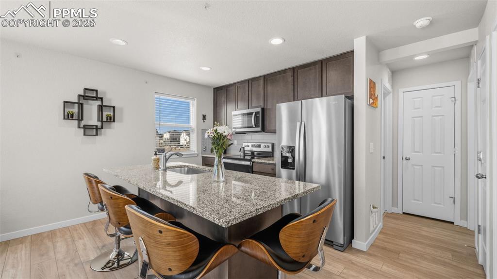Kitchen with dark wood finish cabinetry, stainless steel appliances, a breakfast bar, light stone counters, and a kitchen island with sink