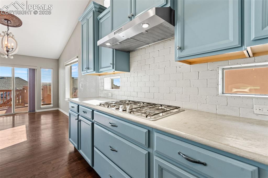 Kitchen with blue cabinets, tasteful backsplash, under cabinet range hood, dark wood-style floors, and vaulted ceiling