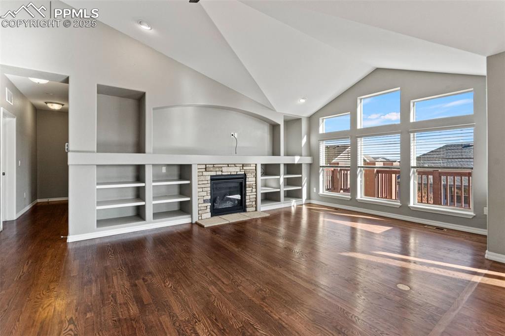 Unfurnished living room with a stone fireplace, dark wood finished floors, built in shelves, high vaulted ceiling, and recessed lighting