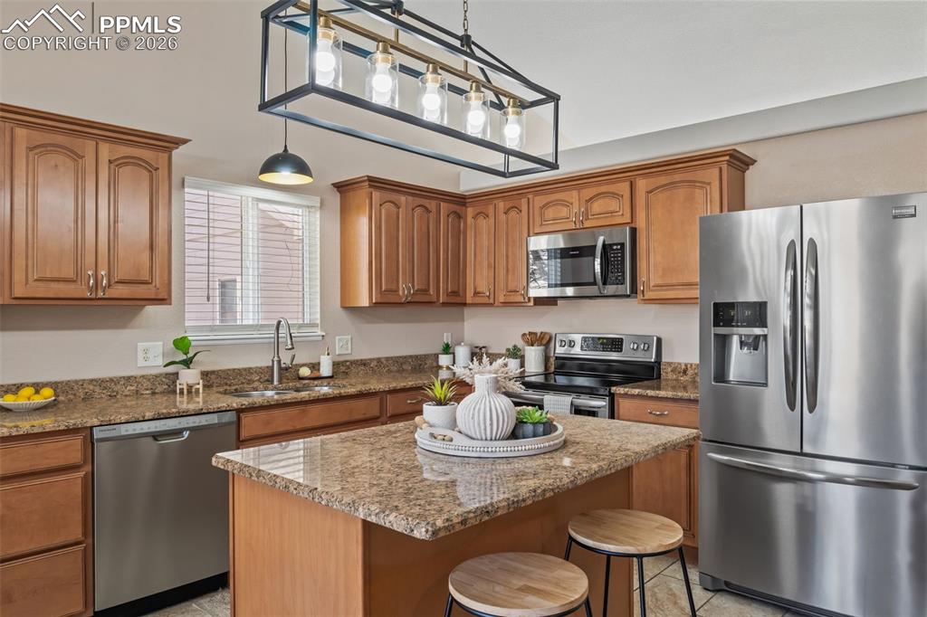 Kitchen with stainless steel appliances, wood finish cabinetry, hanging light fixtures, and light stone counters