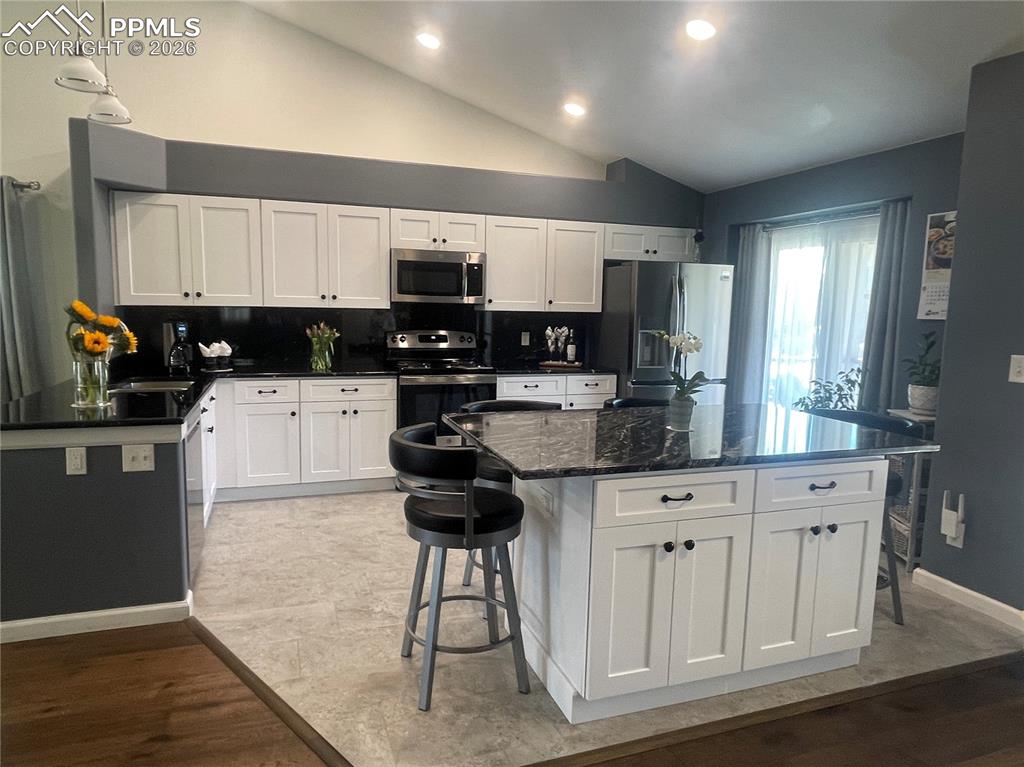 Kitchen with a breakfast bar, vaulted ceiling, white cabinets, stainless steel appliances, and dark stone countertops