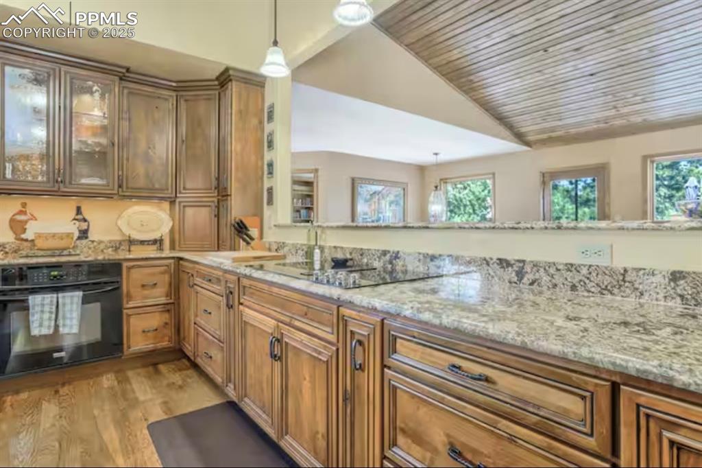 Kitchen featuring brown cabinetry, black appliances, decorative light fixtures, light stone countertops, and lofted ceiling