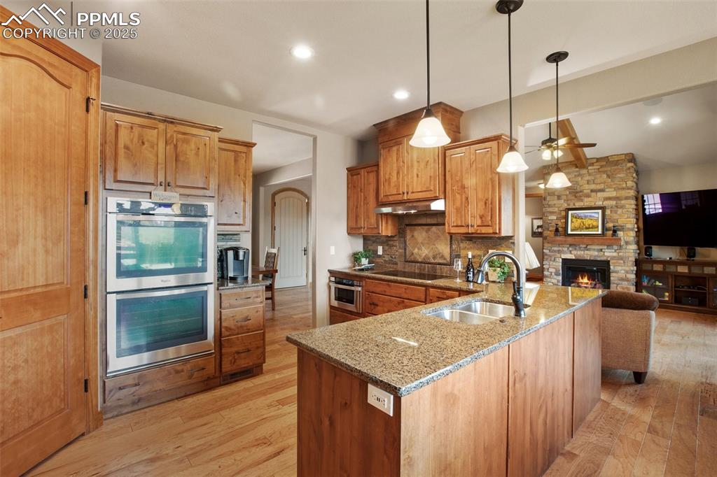 Kitchen featuring black electric stovetop, under cabinet range hood, double oven, a stone fireplace, and a sink