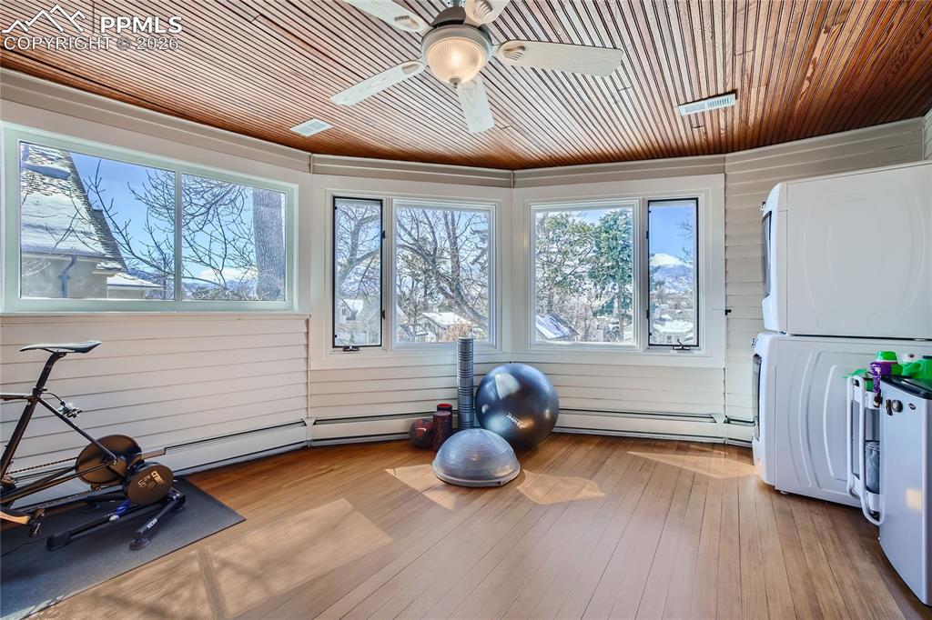 Workout area featuring ceiling fan, a baseboard radiator, wood ceiling, stacked washing maching and dryer, and light hardwood floors