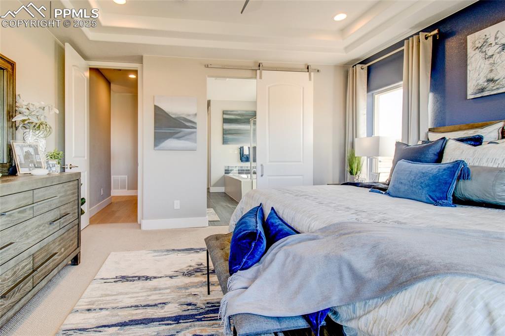 Bedroom featuring a tray ceiling, a barn door, recessed lighting, ensuite bath, and light carpet