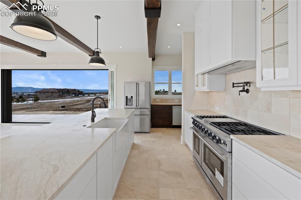 Kitchen with decorative backsplash, beamed ceiling, appliances with stainless steel finishes, glass insert cabinets, and light stone counters