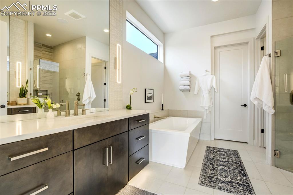 Dual Vanities Finished in Calacatta Lavasa Quartz are Wrapped in Counter-to-Ceiling Tile and Enhanced by Under-Cabinet Lighting and Layered Illumination, Elevating Both Ambiance and Functionality.  At its Center, a Sculptural Freestanding Soaking Tub w/Wall-Mounted Tub Filler Serves as a Striking yet Refined Focal Point—Clean-Lined, Modern + Intentionally Designed.