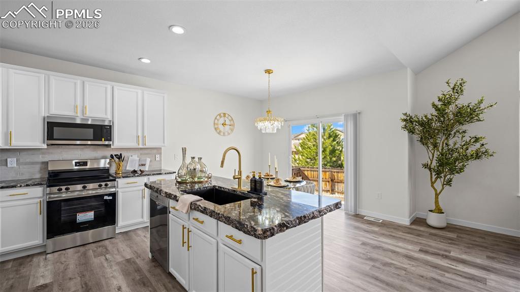 Kitchen with stainless steel appliances, white cabinets, a center island with sink, light wood finished floors, and dark stone countertops