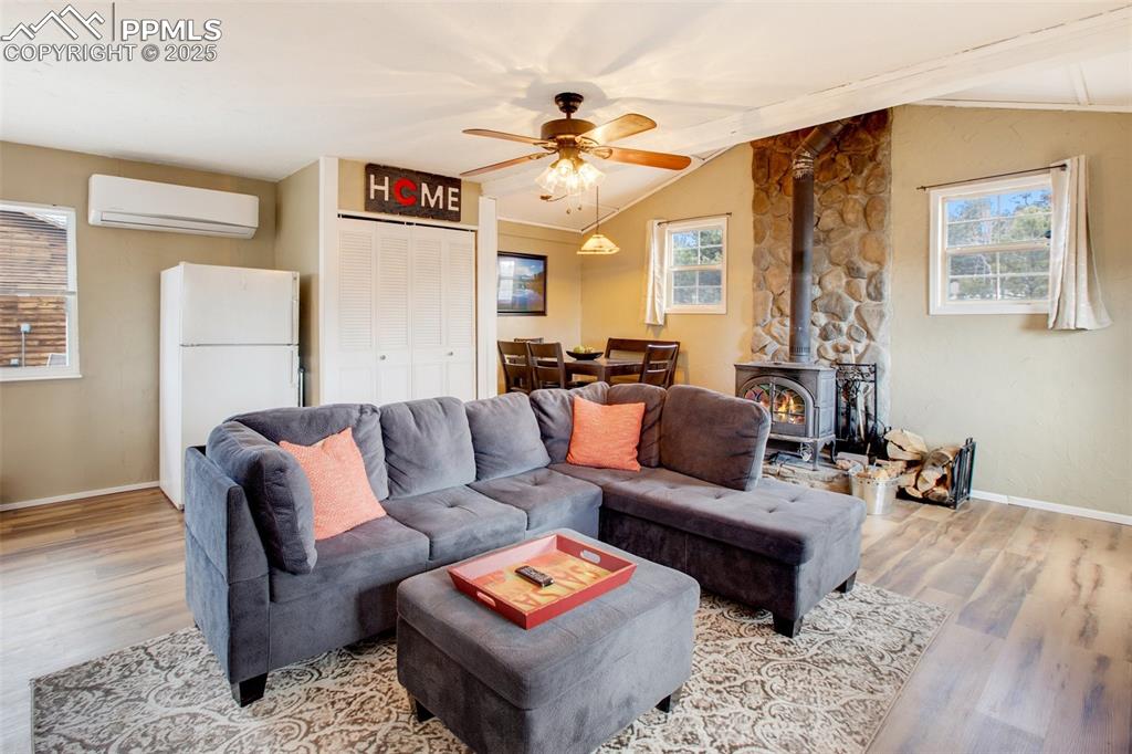 Living room featuring a wood stove, wood finished floors, an AC wall unit, lofted ceiling, and a ceiling fan