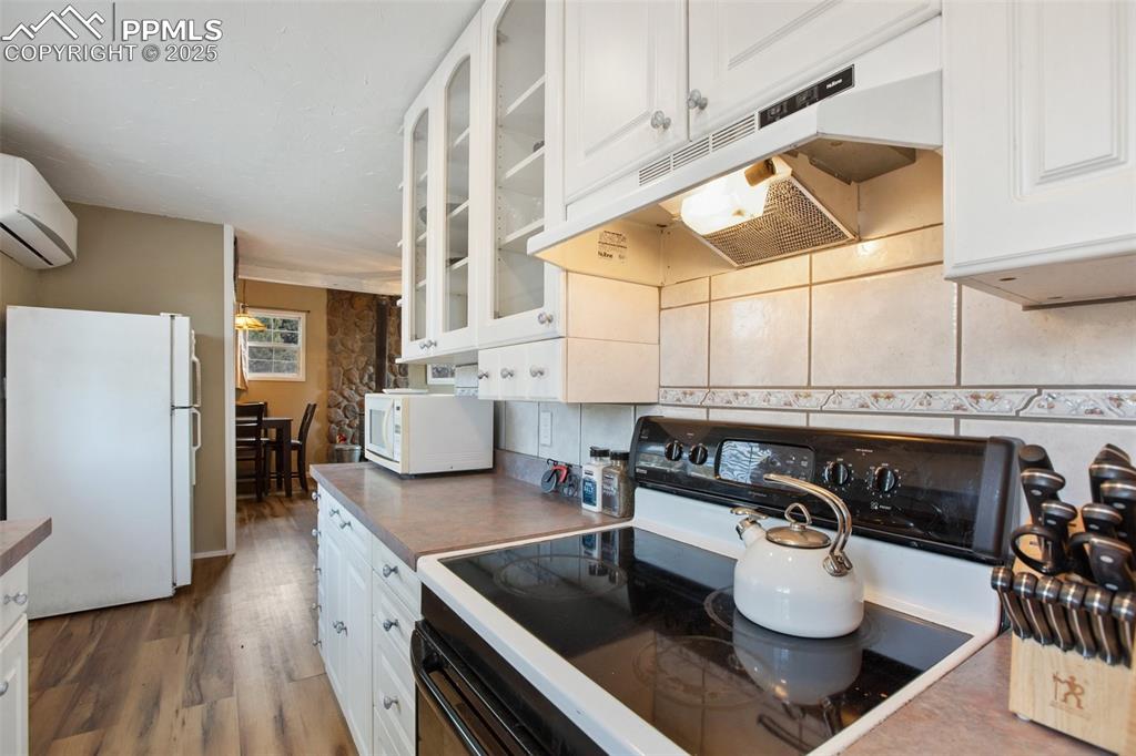 Kitchen with white cabinets, white appliances, under cabinet range hood, wood finished floors, and glass insert cabinets