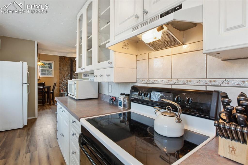 Kitchen with white appliances, white cabinets, under cabinet range hood, wood finished floors, and glass insert cabinets