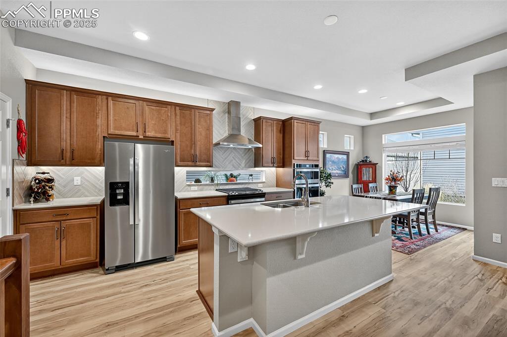Kitchen featuring appliances with stainless steel finishes, decorative backsplash, brown cabinets, wall chimney range hood, and recessed lighting