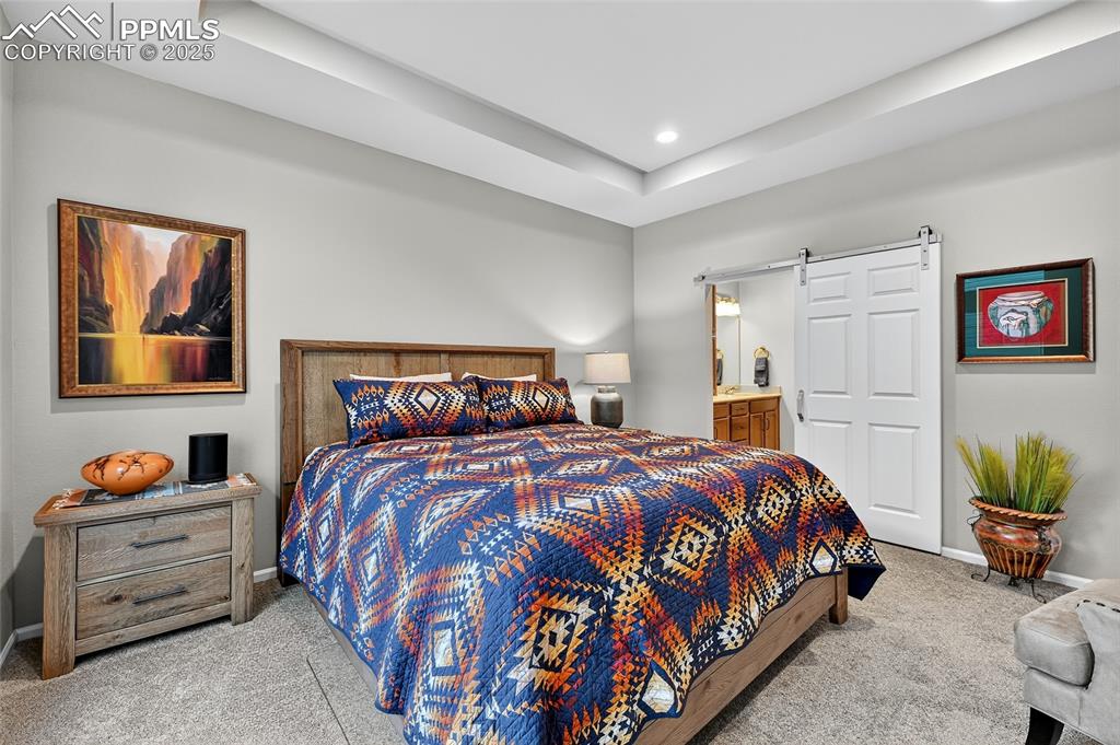Bedroom featuring light colored carpet, a barn door, ensuite bath, recessed lighting, and a tray ceiling