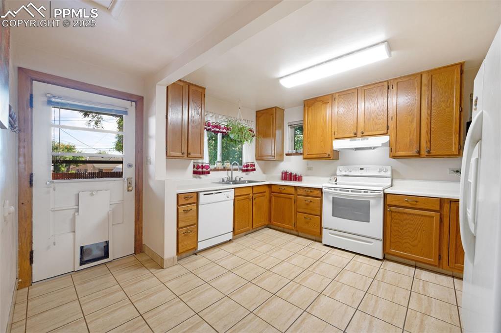 Kitchen with white appliances, light countertops, light tile patterned flooring, brown cabinets, and under cabinet range hood