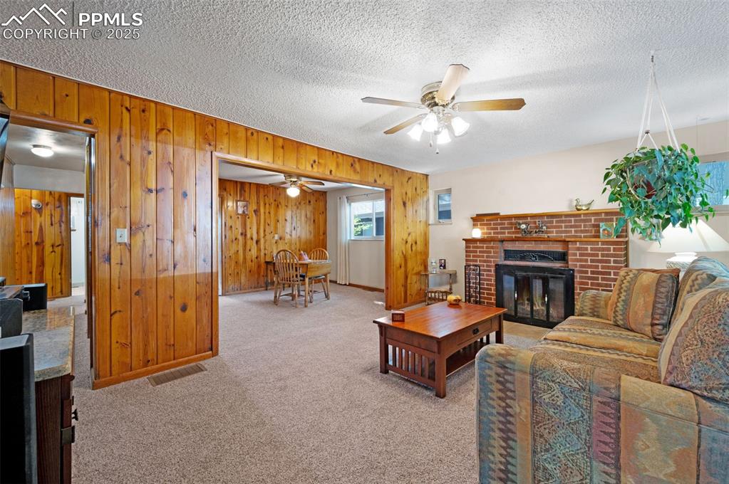 Carpeted living area featuring a brick fireplace, wood walls, a textured ceiling, and a ceiling fan