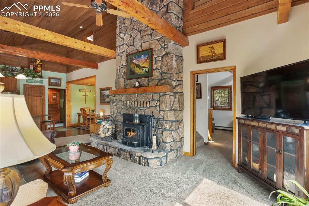 Living room featuring carpet flooring, a wood ceiling with exposed beams, a wood stove, and a ceiling fan!