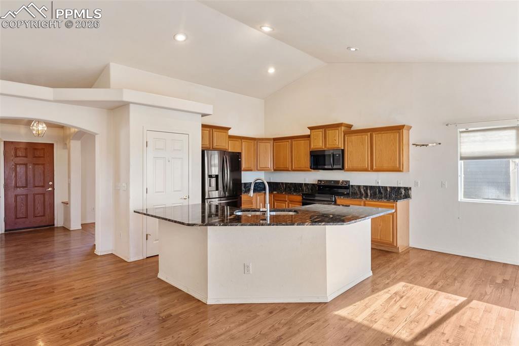 Kitchen with an island with sink, black appliances, dark stone countertops, arched walkways, and light wood-style floors