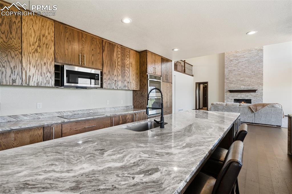 Kitchen featuring appliances with stainless steel finishes, brown cabinetry, light stone counters, dark wood finished floors, and a stone fireplace